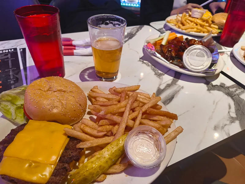 アメリカのハンバーガー。上のバンズは外されており、チーズが載せられたパティが見える。太めのフライドポテトと切られていないピクルス、透明なプラカップに入れられた白いソースが皿に載っている。机の上には若干飲まれたビールと、赤いコップに入れられた水が見える。また、奥にはバッファローチキンの皿があり、同じく透明なプラカップに入った白いソースもある。