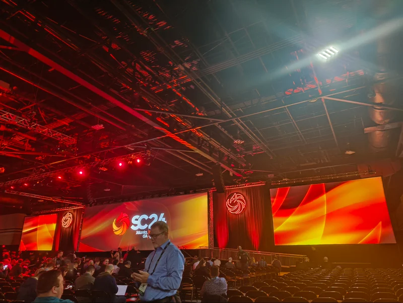 SC24のオープニングの画像。巨大な三枚のスクリーンを使いSCのロゴが映し出されていrう。照明により、SC24のテーマカラーである赤色で会場が染まっている