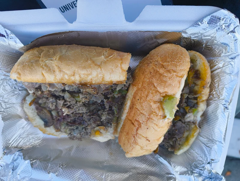 Philly steaks。表面が少しカサカサしてそうなコッペパンのようなパンが割られ、中に肉が詰まっている。パンは20 cmくらいありそうだが、半分で切られて容器に収まっている。